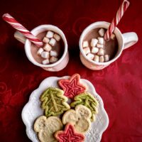two cups of cocoa with peppermint sticks and mini marshmallows and a plate of christmas cookies