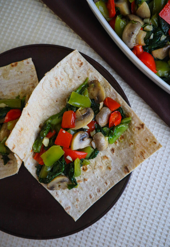 an all vegetable saute for wraps with escarole green and red peppers mushrooms and garlic on a wrap served on brown plate