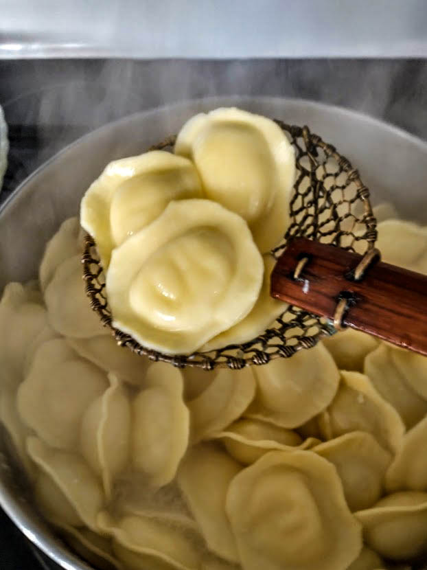  4 round cheese filled raviolis on a straining spoon over a pot of slow boiling water with the rest of the ravioli cooking